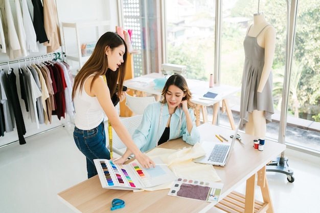 Duas estilistas ou costureiras asiáticas debateram e discutiram o design da nova coleção com exemplos de tecidos e esboços no tablet Trabalhando juntas no estúdio