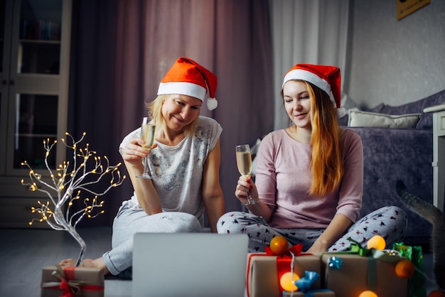 Duas feliz animada jovem com chapéus de Papai Noel brindando com vinho espumante na webcam do laptop. Casal feminino do amor fazendo videochamada de ano novo em casa.