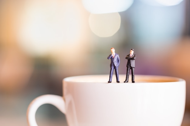 Duas figuras diminutas do homem de negócios estão e pensando na placa branca do copo do café quente.