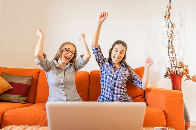 Duas garota muito feliz torcendo com as mãos para cima, olhando para a tela do laptop, sentado no sofá em casa. mulheres jovens vencedoras tendo sucesso em seu trabalho. mude sua vida e faça o trabalho preferido para viver melhor