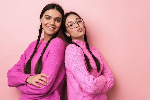 Duas garotas adolescentes alegres e fofas em pé, costas com costas, isoladas sobre uma parede rosa