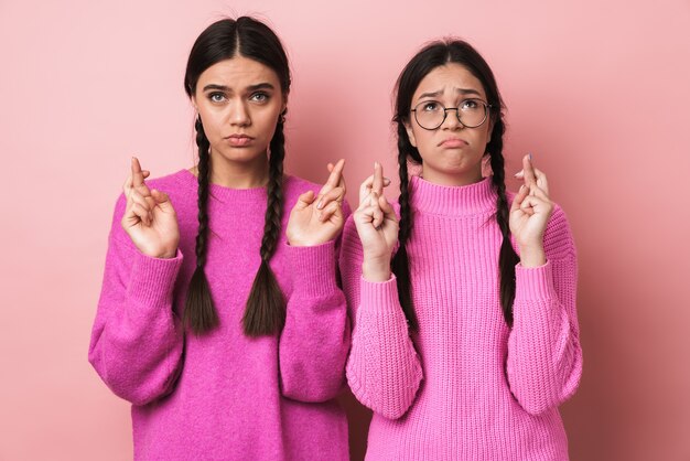 Duas garotas adolescentes fofas e chateadas de pé isoladas sobre uma parede rosa, cruzando os dedos para dar sorte