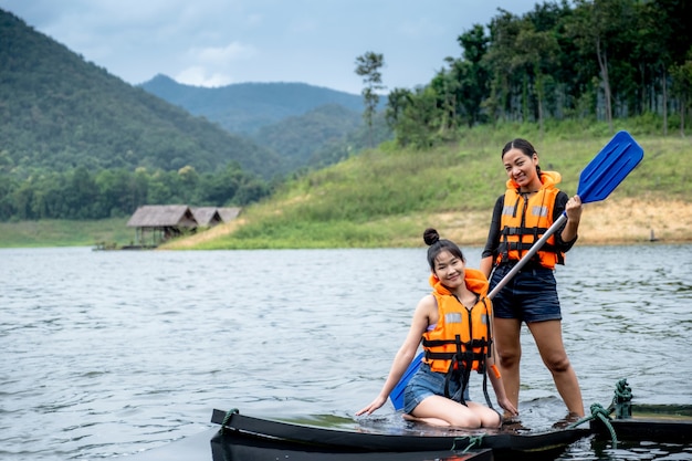 Duas garotas asiáticas em coletes salva-vidas laranja uma pessoa segura um remo, a outra senta-se, tendo como pano de fundo água e montanhas pronta para viajar como hobby.