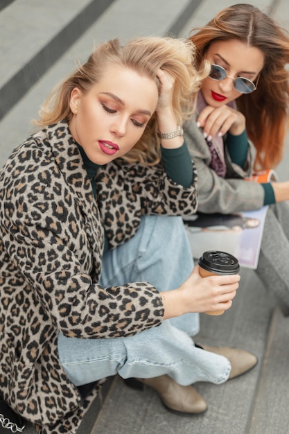 Duas garotas bonitas da moda loiras e ruivas em roupas casuais da moda sentadas nos degraus da cidade e bebendo café