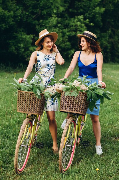 Duas garotas bonitas vão com bicicletas no parque primavera ao pôr do sol. Namoradas se divertem, riem, sorriem e andam de bicicleta. Retrato de corpo inteiro de duas jovens mulheres em vestidos e chapéus de palha. feriado ativo