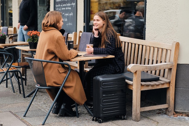 Duas garotas casuais atraentes conversando alegremente durante a pausa para o café com mala no café de rua