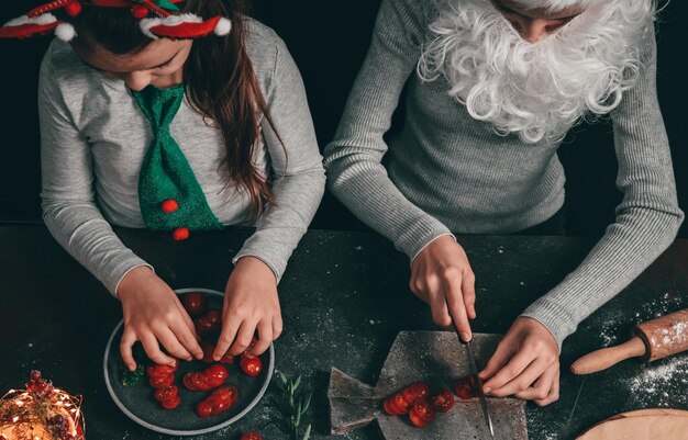 Duas garotas caucasianas com chapéu de Papai Noel com barba e bandana estão felizes sentadas à mesa e preparando pizza com ingredientes