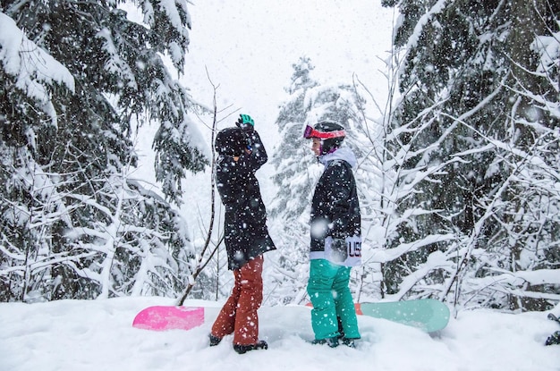 Duas garotas com pranchas de snowboard na floresta nas montanhas e a queda de neve