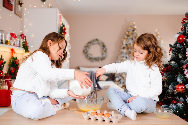 Duas garotas cozinham bolo de Natal sentadas na mesa da cozinha sobre as decorações de Natal no quarto em casa Temporada de férias de inverno Infância