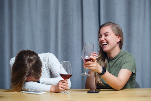 Duas garotas de aparência europeia com cabelos loiros estão sentadas à mesa, bebendo vinho e rindo, relaxando em casa, álcool