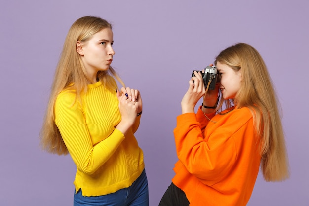 Duas garotas de irmãs gêmeas loiras jovens encantadoras com roupas coloridas, tirando fotos na câmera fotográfica vintage retrô isolada na parede azul violeta. conceito de estilo de vida familiar de pessoas.