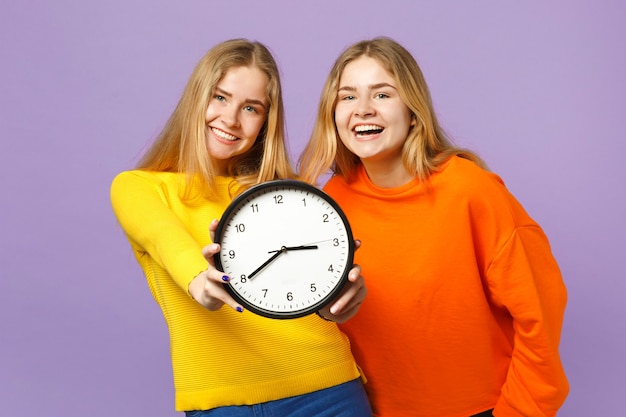 Duas garotas de irmãs gêmeas loiras jovens rindo em roupas coloridas vivas segurar o relógio redondo isolado na parede de azul violeta pastel. Conceito de estilo de vida familiar de pessoas.