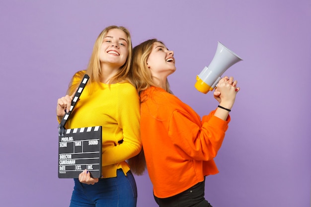 Duas garotas de irmãs gêmeas loiras jovens rindo segurar clássico filme preto fazendo claquete, gritar no megafone isolado na parede azul violeta. Conceito de estilo de vida familiar de pessoas.