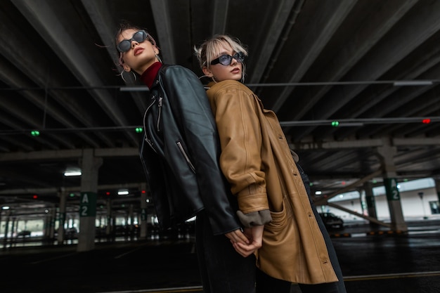 Duas garotas de moda em roupas elegantes da cidade, jaqueta de couro e óculos de sol legais, posando perto de um estacionamento na cidade