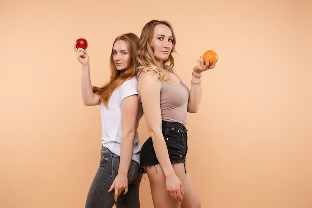 Duas garotas desportivas, olhando para a câmera e posando com frutas e vegetais úteis em fundo isolado. irmãs jovens em pé, costas com costas, mantendo a maçã e a laranja e rindo.