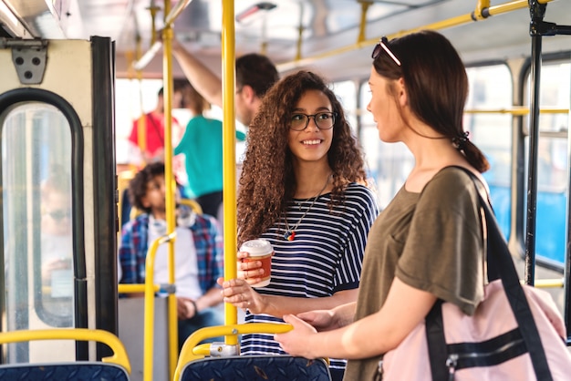 Foto duas garotas em pé em um ônibus conversando e tomando café.