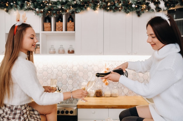 Duas garotas em um ambiente familiar aconchegante na cozinha servem champanhe para o natal. garotas sorridentes bebem champanhe em uma noite festiva