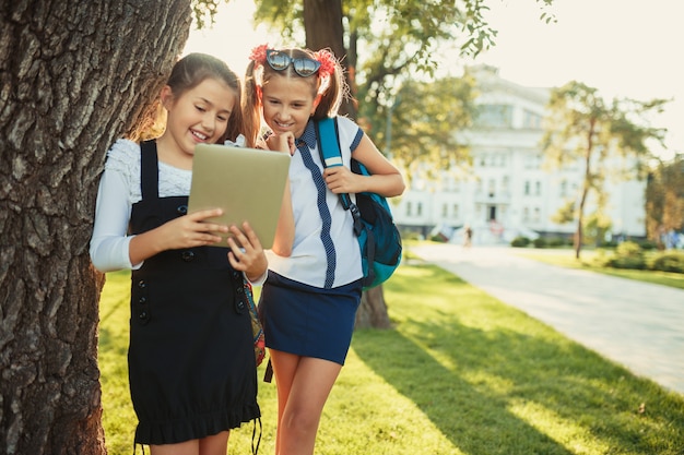 Duas garotas encantadoras em idade escolar ficam perto da árvore e brincam no tablet.
