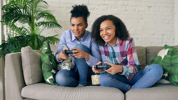 Duas garotas encaracoladas sentadas no sofá jogam jogos de computador de console com gamepad e se divertem em casa