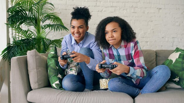 Foto duas garotas encaracoladas sentadas no sofá jogam jogos de computador de console com gamepad e se divertem em casa