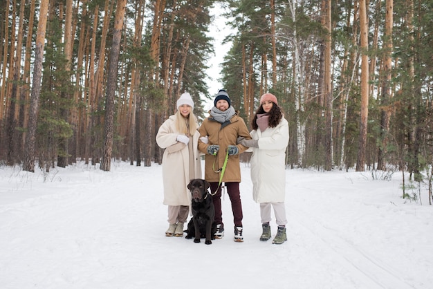 Duas garotas felizes em roupas de inverno ao lado de um jovem com um retriever preto enquanto caminhava em uma floresta ou parque coberto de neve