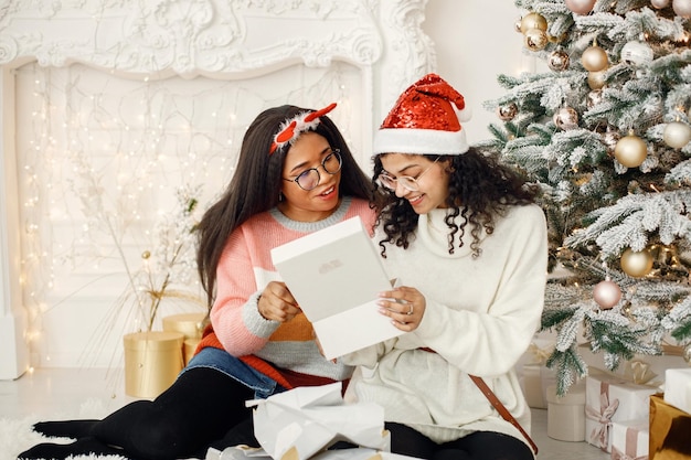 Duas garotas indianas em óculos abrindo um presente perto da árvore de Natal