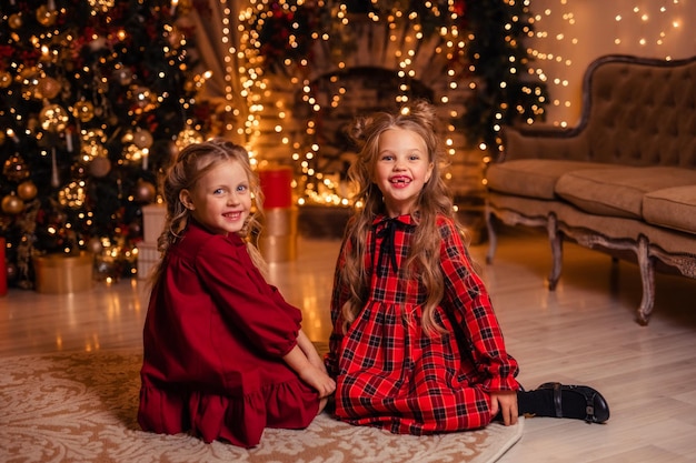 Duas garotas lindas e alegres em vestidos vermelhos brincando ao lado da árvore de Natal no fundo da lareira