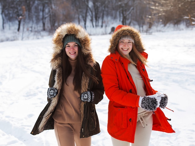 Duas garotas no inverno, com roupas quentes, curtindo a neve, ao ar livre. Época fria do ano, risos e diversão. Floresta de inverno, clareira branca.