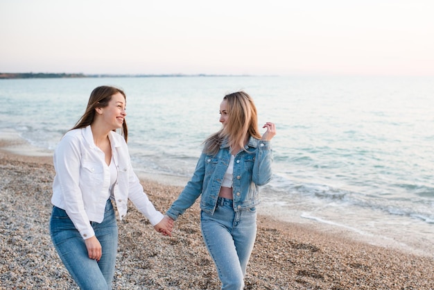 Duas garotas rindo felizes usam roupas jeans se divertindo andando na praia arenosa sobre o mar