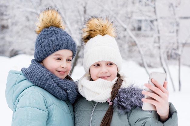 Duas garotas sorridentes em casacos e chapéus quentes são fotografadas em um smartphone em um parque de inverno nevado Uso de tecnologia de estilo de vida