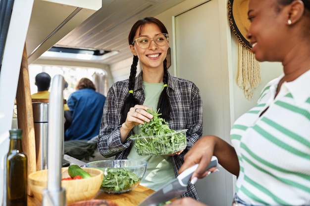 Duas garotas sorrindo uma para a outra enquanto cozinhavam salada de legumes juntas no motorhome durante o tra