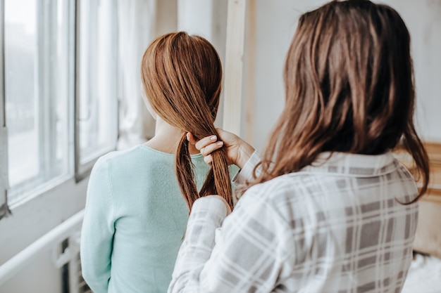 Duas garotas trançam os cabelos na janela. mulher faz uma trança para a amiga. penteados de tecelagem de cabelo. namorada trança as mãos com cachos. cuidado capilar