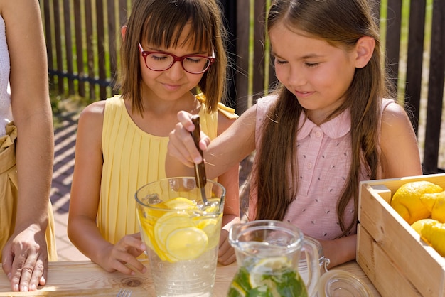 Duas garotinhas bonitas ajudando a mãe a fazer limonada fresca para vender, enquanto uma delas espreme uma rodela de limão na jarra com água