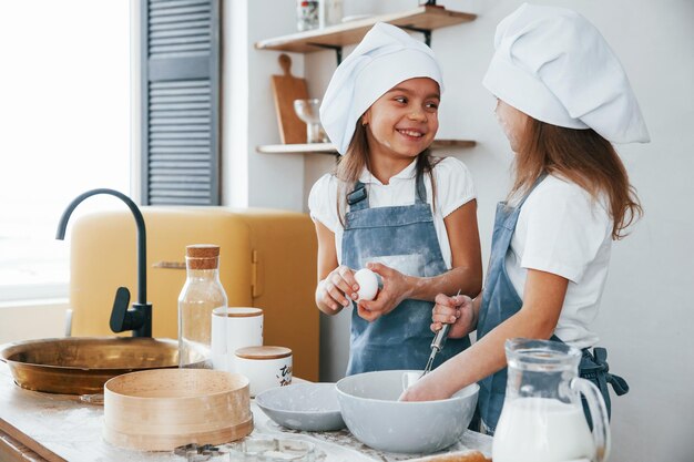 Duas garotinhas de uniforme de chef azul conversando segredos umas com as outras ao preparar comida na cozinha