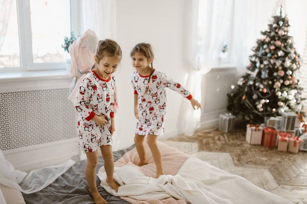 Duas garotinhas encantadoras de pijama se divertem pulando em uma cama em um quarto aconchegante e iluminado pelo sol, com árvore de ano novo e presentes. .