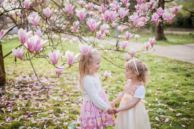 Duas garotinhas loiras de 3 anos de idade estão brincando no parque perto de uma magnólia em flor. Beber chá. Páscoa. Primavera.