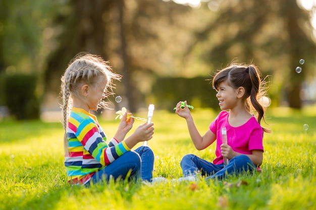 Duas garotinhas loiras e morena de verão sentadas no gramado soprando bolhas de sabão Crianças de etnia europeia e indiana
