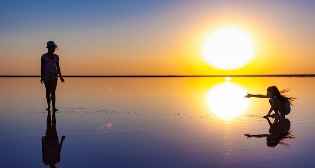 Duas irmãs adoráveis e felizes estão caminhando ao longo do lago salgado espelhado apreciando o pôr do sol ardente