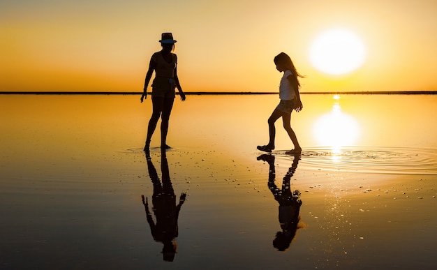 Duas irmãs adoráveis e felizes estão caminhando ao longo do lago salgado espelhado apreciando o pôr do sol ardente