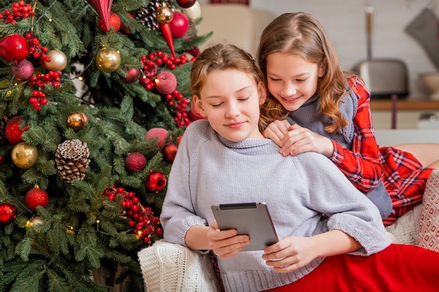 Duas irmãs alegres estão sentadas em casa perto da árvore de Natal e olhando para o monitor do tablet