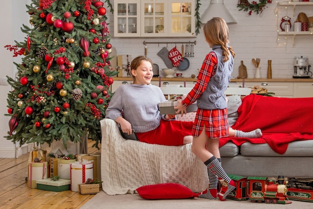 Duas irmãs alegres estão sentadas em casa perto da árvore de Natal e se cumprimentam no Natal, dando presentes.