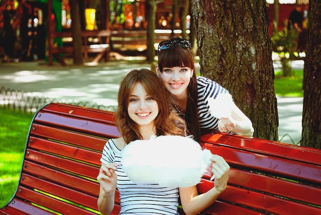 Duas irmãs, amigas se divertem, riem, comem algodão doce, caminham em um parque de diversões verde de verão. Feliz