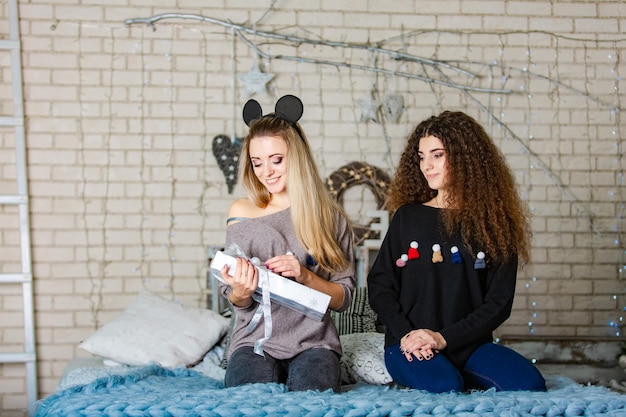 Duas irmãs bonitas em suéteres de Natal sentadas de joelhos em uma manta de malha e segurando um presente de Ano Novo enquanto passam tempo juntos