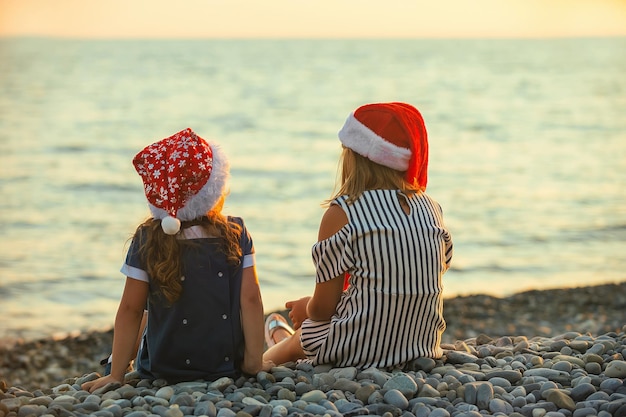 Duas irmãs com chapéus de papai noel sentam e conversam alegremente à beira-mar ao pôr do sol