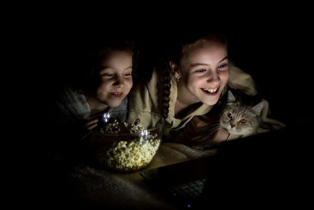 Foto duas irmãs com um gato assistem a desenhos animados no dispositivo e se divertem juntas lazer da infância