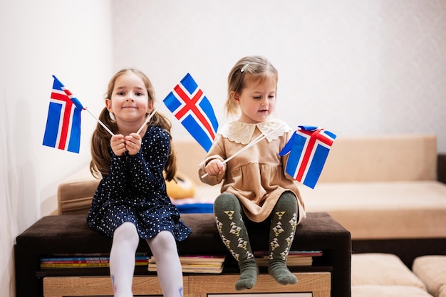 Duas irmãs estão sentadas em um sofá em casa com bandeiras islandesas nas mãos islândia crianças meninas com bandeira