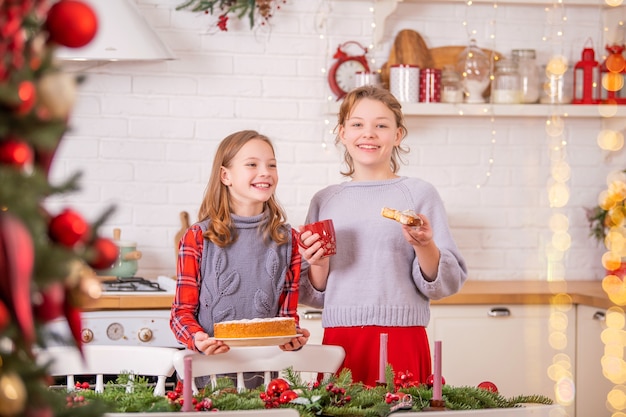 Duas irmãs felizes estão decorando a mesa de Natal da cozinha, segurando xícaras e tortas doces nas mãos.