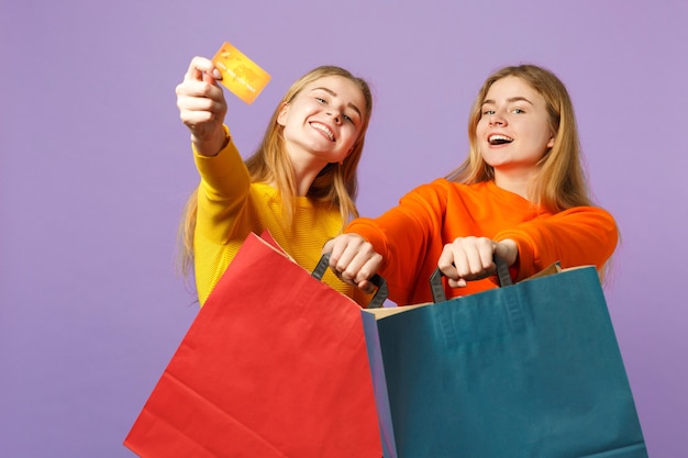 Duas irmãs gêmeas loiras engraçadas com roupas vivas, segurando o cartão do banco de crédito, sacola de pacote com compras depois de fazer compras isoladas na parede azul violeta. conceito de família de pessoas.
