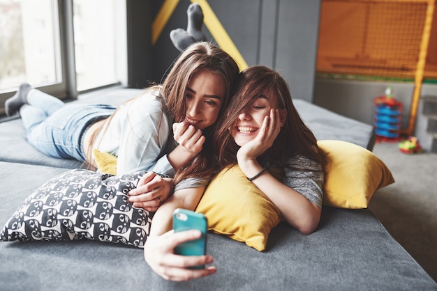 Duas irmãs gêmeas sorridente fofo segurando o smartphone e fazendo selfie. Meninas mentem no sofá posando e alegria