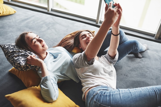 Duas irmãs gêmeas sorridente fofo segurando o smartphone e fazendo selfie. Meninas mentem no sofá posando e alegria